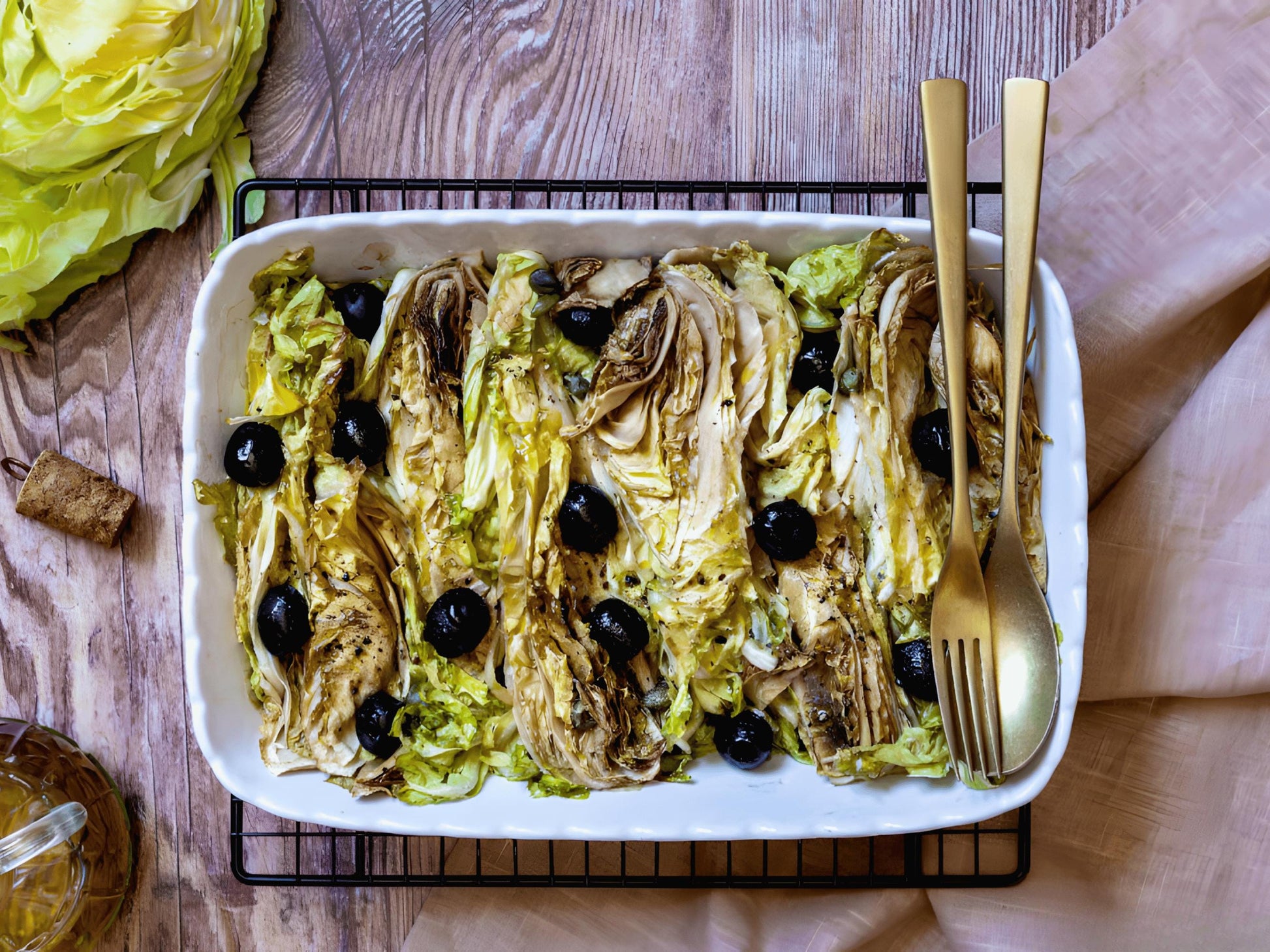 A white ceramic dish filled with a salad of lettuce, black olives, and other vegetables, accompanied by a fork and knife on a wooden table.
