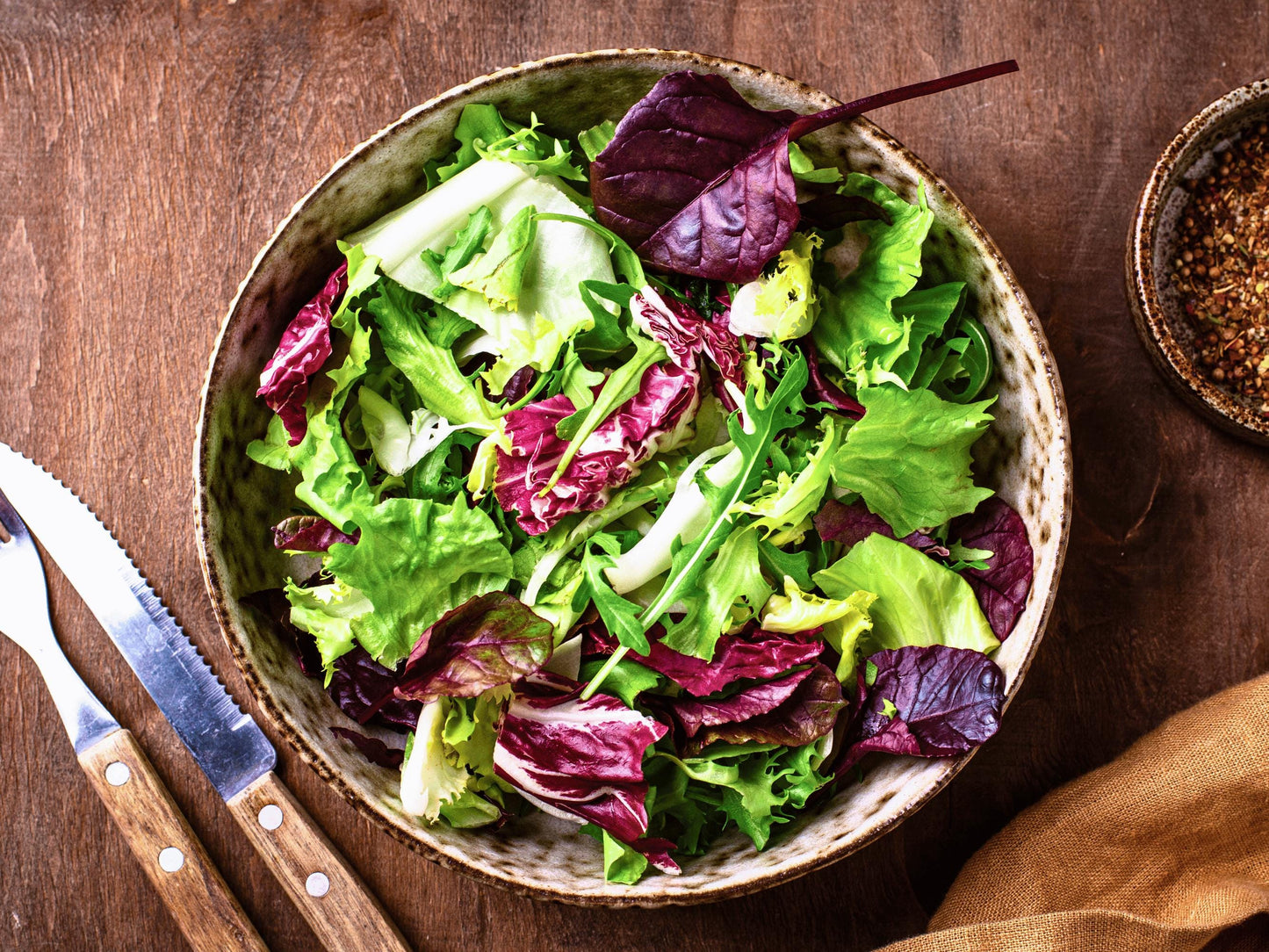 A bowl of fresh salad with various green and purple lettuce leaves, accompanied by a knife and a small bowl of seasoning on a wooden table.