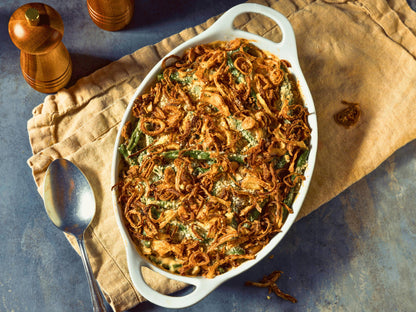 A white ceramic dish filled with a green bean casserole, topped with fried onions, sits on a beige cloth napkin. A wooden pepper grinder and a spoon are placed next to the dish, and a wooden cutting board is visible in the background.