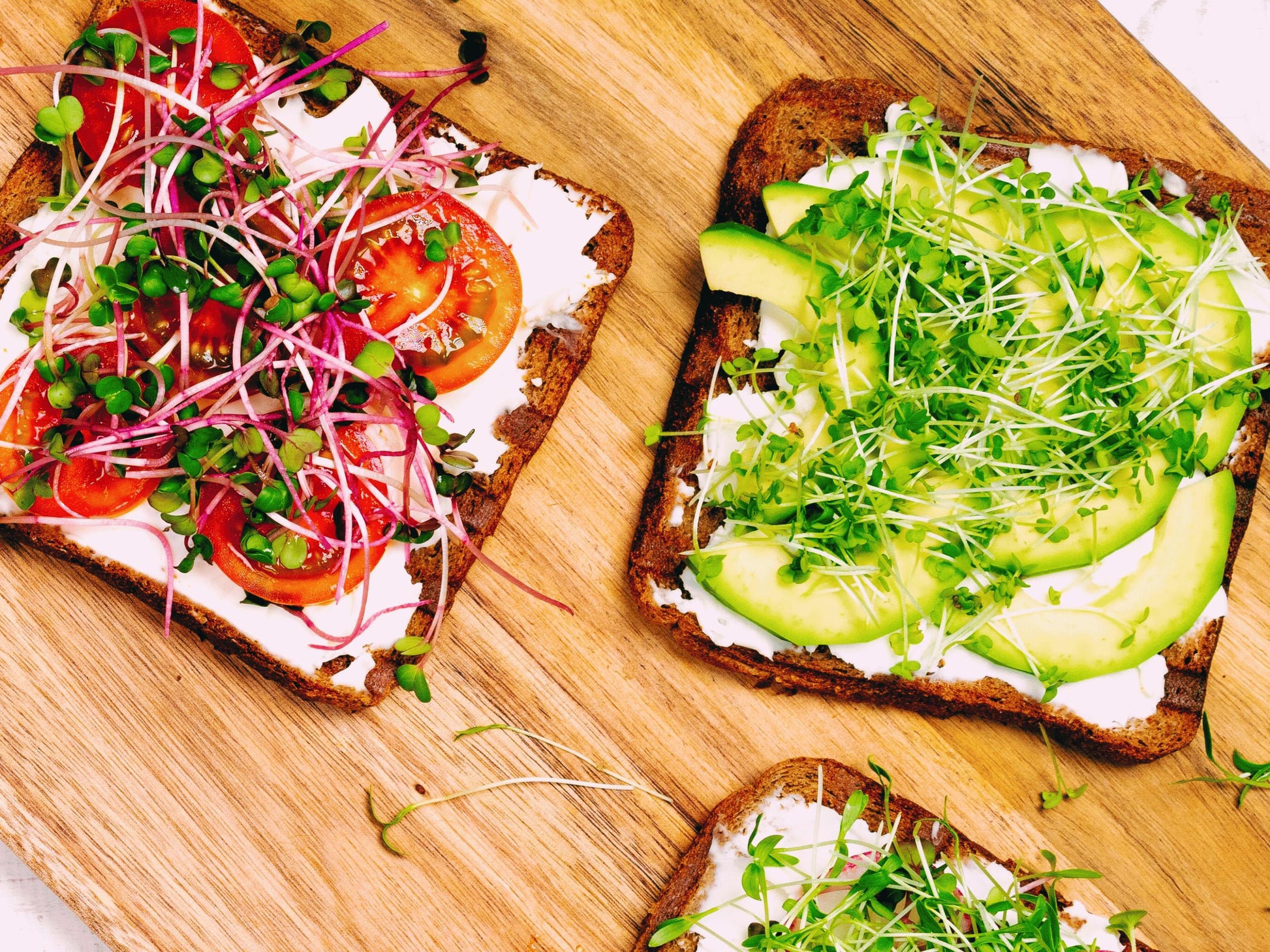 three slices of bread topped with various fresh ingredients, including avocado, tomatoes, and sprouts, arranged on a wooden surface.