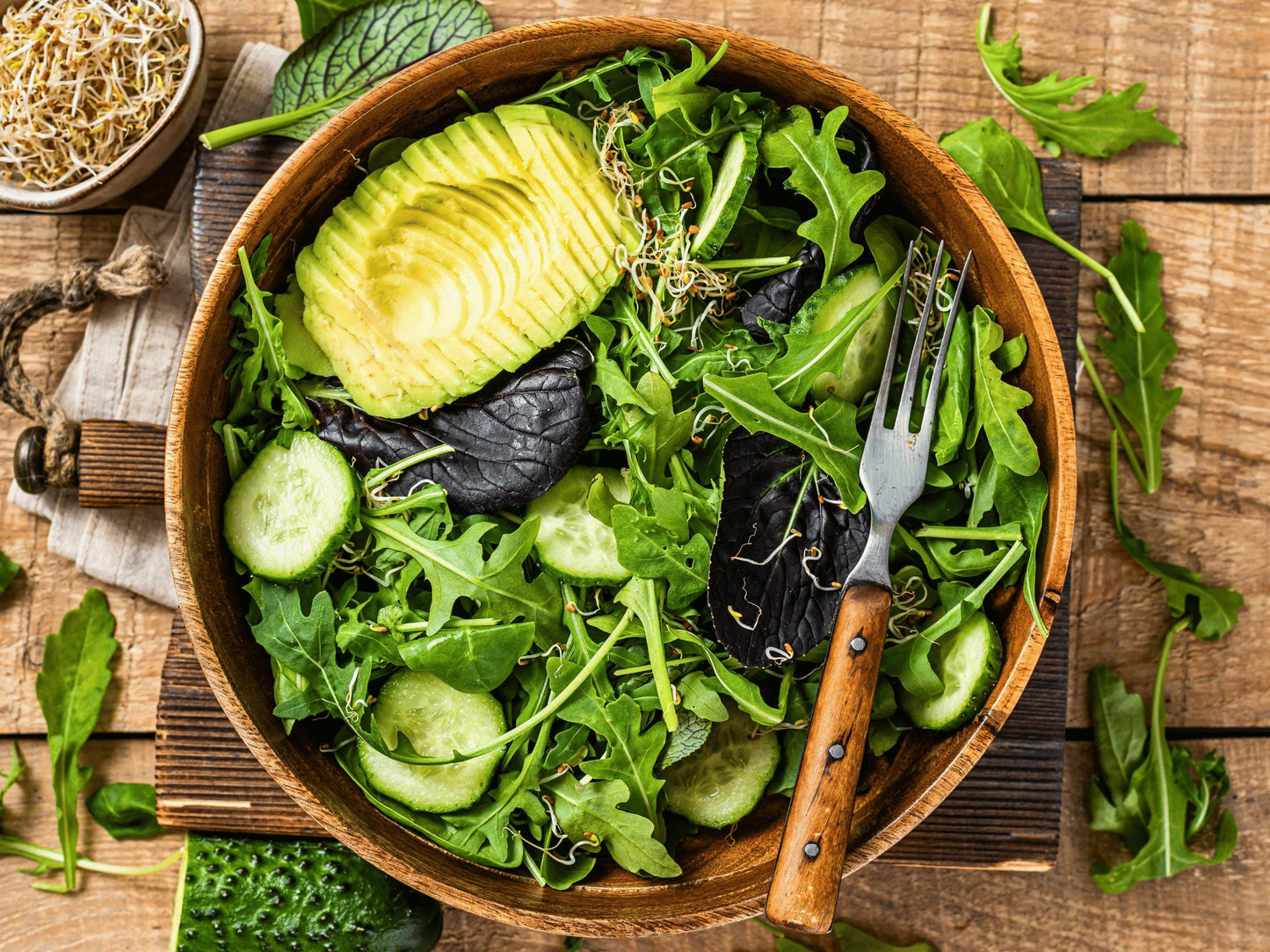 A wooden bowl filled with a fresh salad of various greens, cucumbers, and avocado slices, accompanied by a fork on a wooden surface.