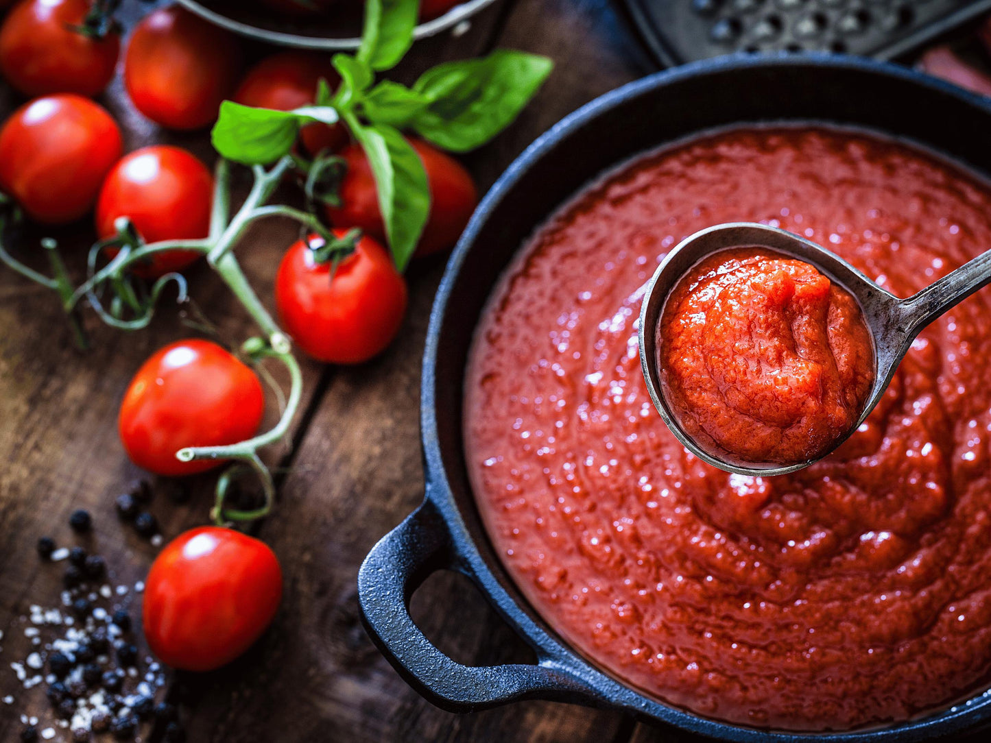 A bowl of vibrant red tomato sauce with a ladle, surrounded by fresh tomatoes, basil leaves, and a black pan.