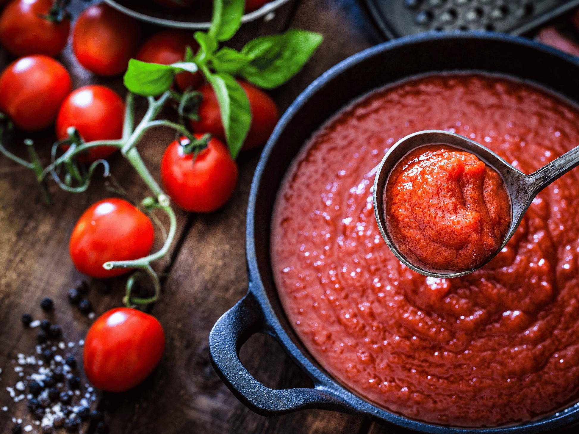 A bowl of vibrant red tomato sauce with a ladle, surrounded by fresh tomatoes, basil leaves, and a black pan.