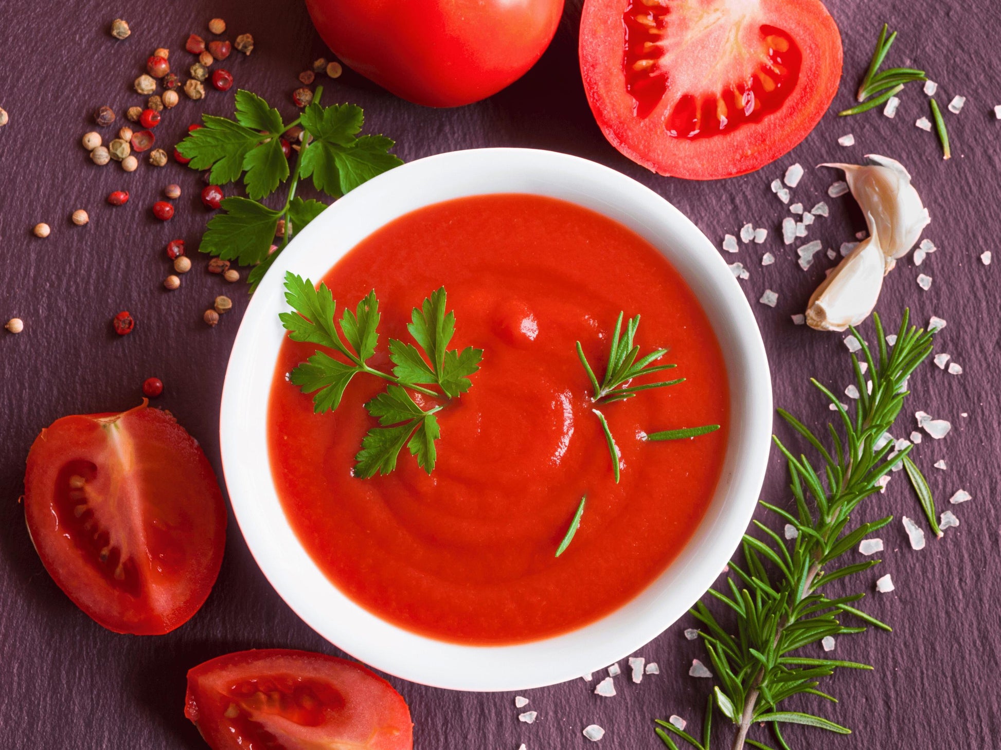 A bowl of vibrant red tomato soup is surrounded by fresh herbs, spices, and sliced tomatoes on a dark purple background.
